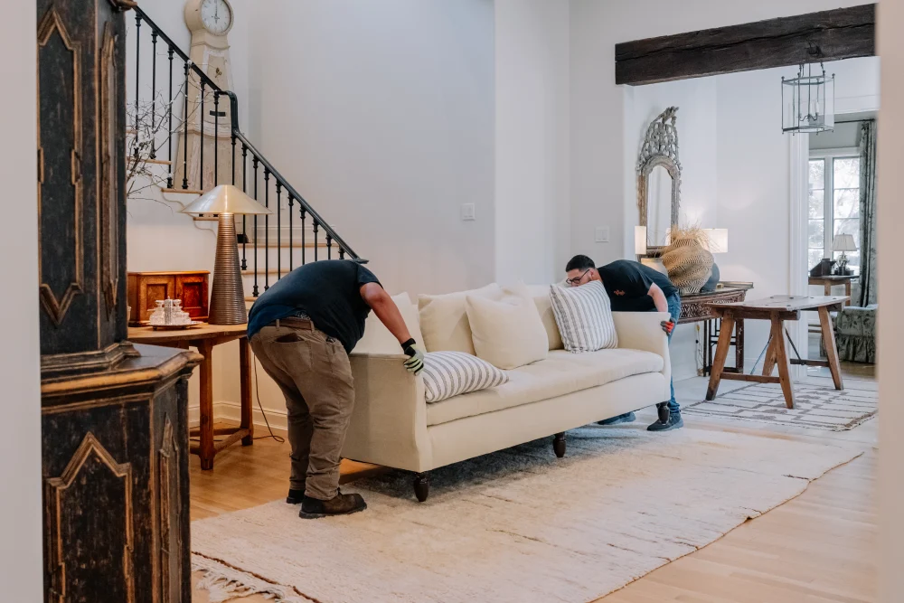 Two movers carefully carry a cream sofa into a stylish home with a grand staircase.