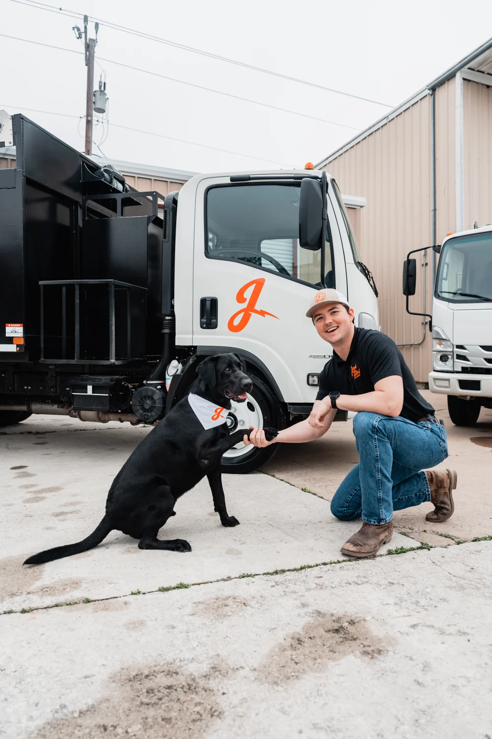 A smiling man kneels, shaking paws with a black dog in front of a white