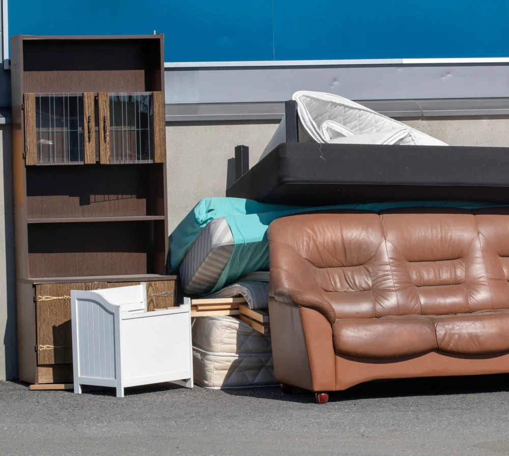 Discarded brown leather sofa, bookshelf, and mattresses piled outdoors for bulky waste collection
