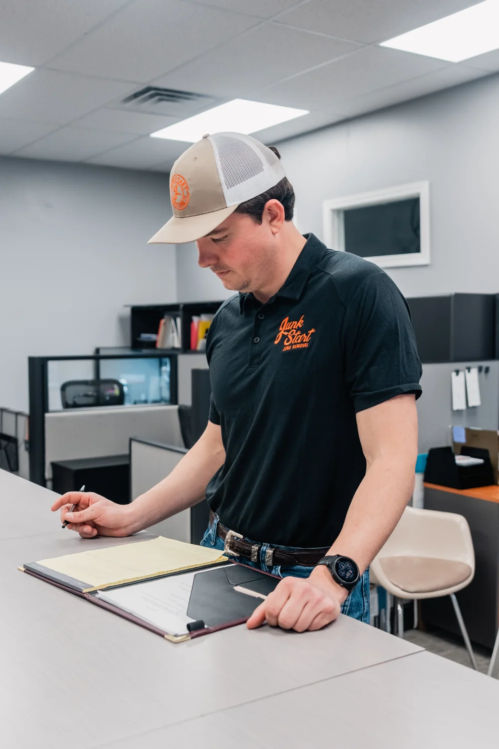A man in a Junk Start uniform cap and shirt reviews paperwork at an office counter.