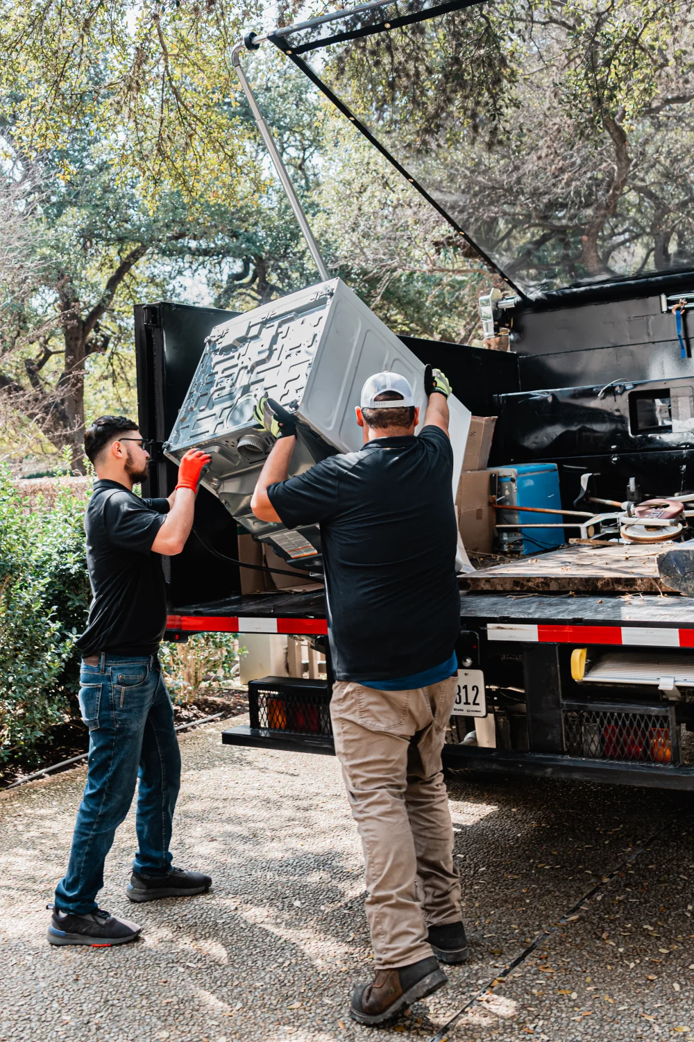 Two men wearing work gloves load a large white appliance into a black junk removal truck.