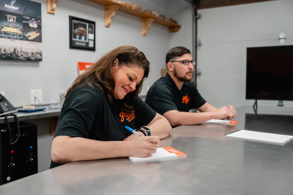 A smiling woman in a "Junk Start" shirt writes at a table with a man beside her.