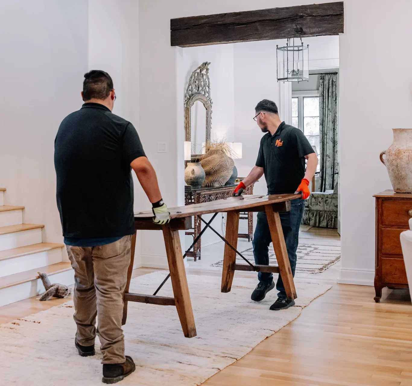 Professional movers carefully carry a rustic wooden table through a bright, elegant home.