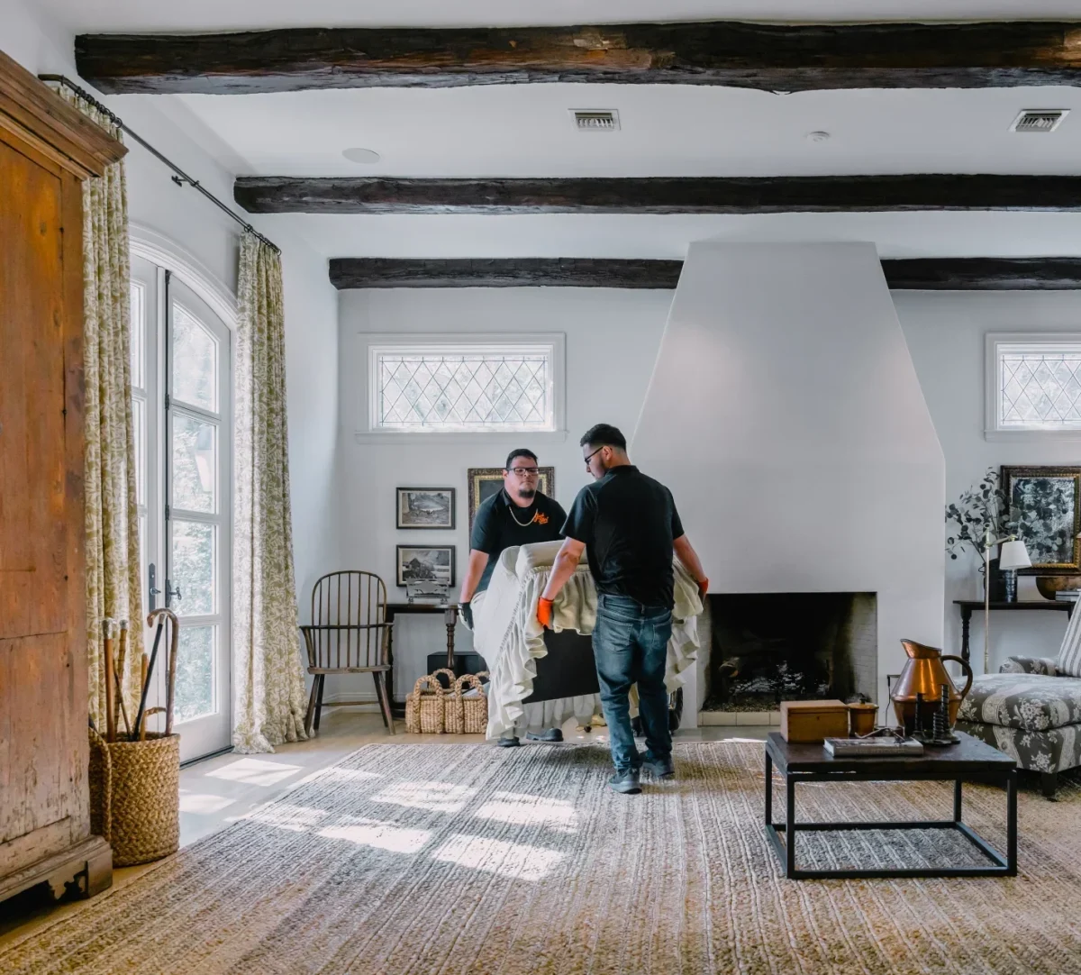 Two movers carry furniture in a bright, rustic living room with exposed beams and a large fireplace