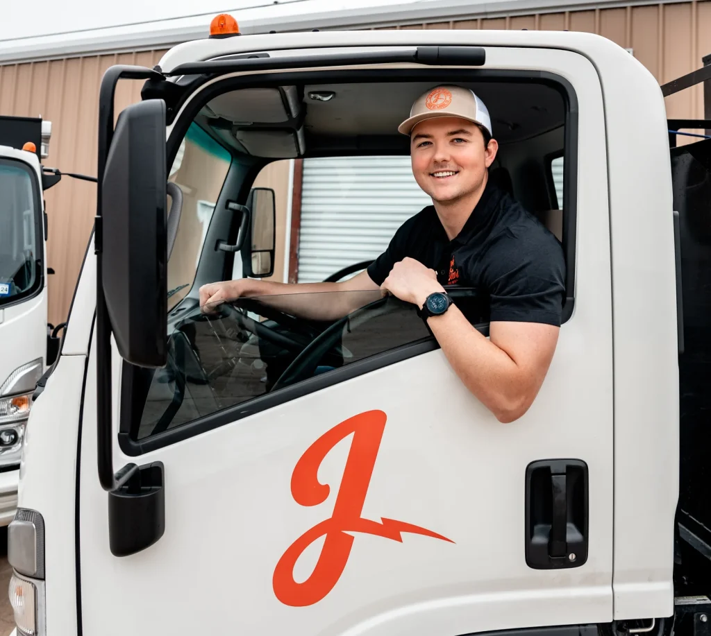 A friendly young man in a cap smiles from a white service truck featuring an orange 'J