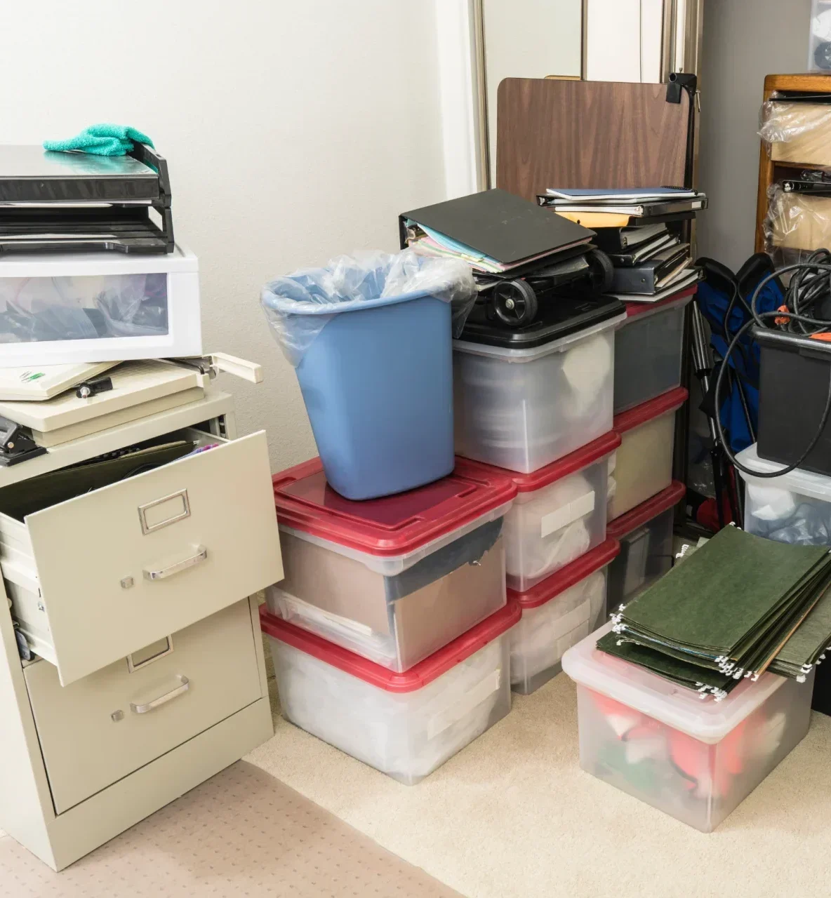 A cluttered room filled with stacks of storage bins, a filing cabinet, and various office