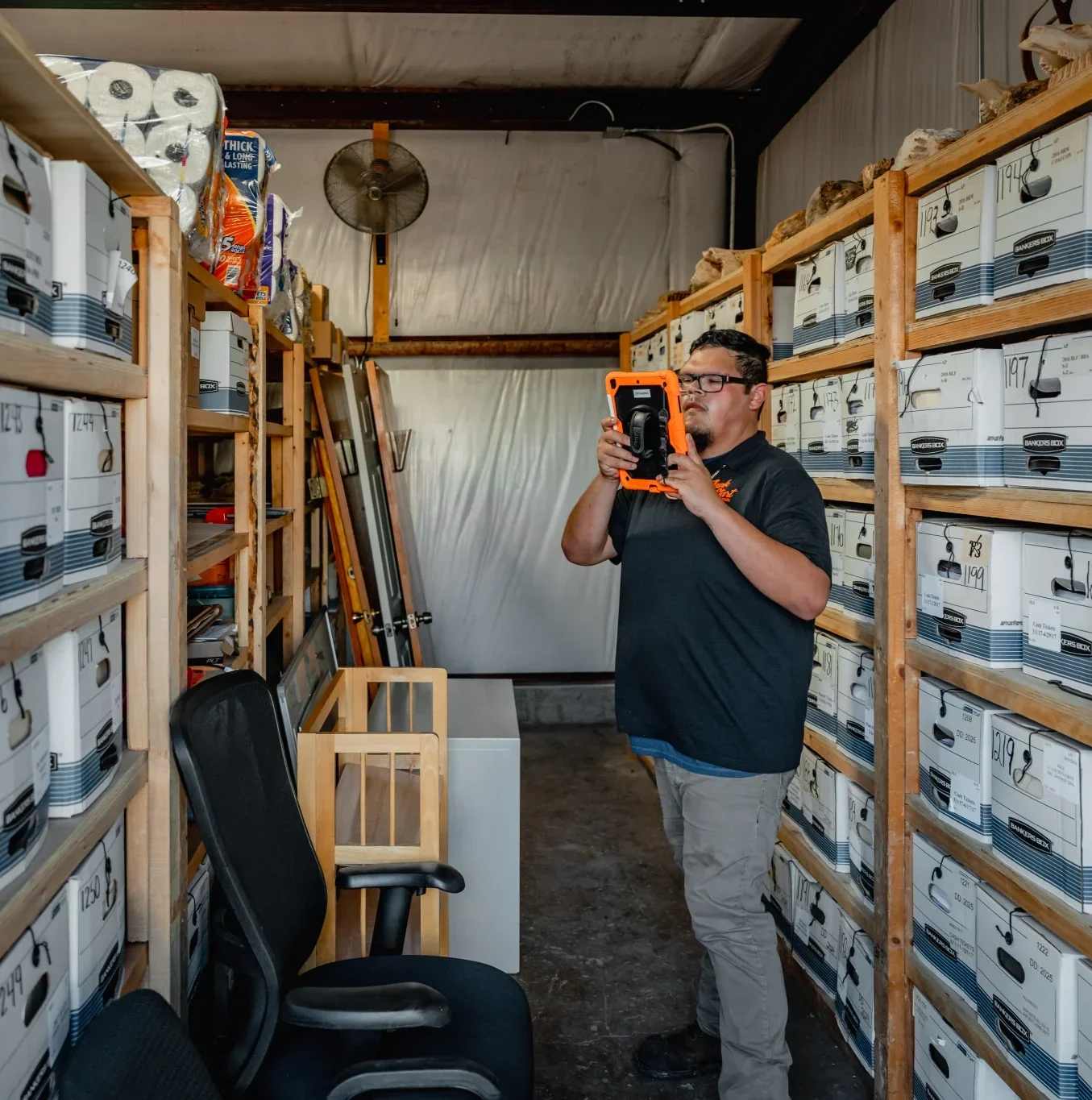 A man scans white storage boxes on wooden shelves in a cluttered archive room using an