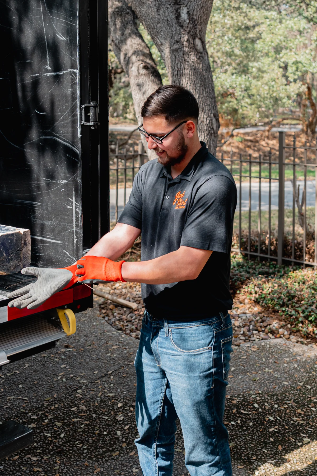 A man wearing a Junk Start shirt and glasses puts on work gloves by a junk removal truck.