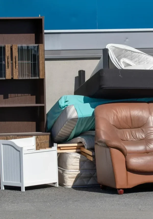 A brown leather sofa, wooden bookshelf, and mattresses are piled outdoors for disposal. TITLE: