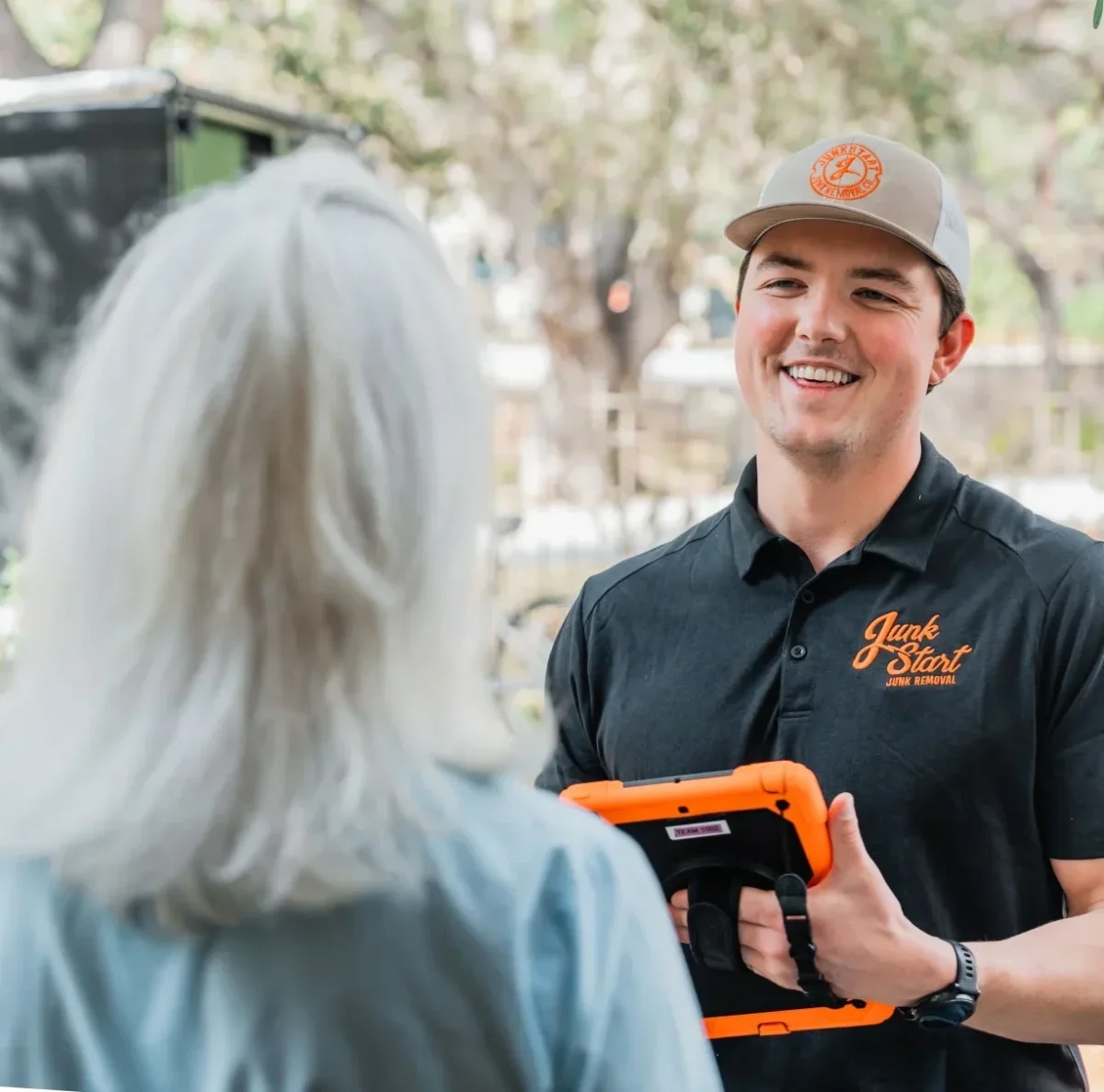 A smiling Junk Start employee talks to a customer at their doorstep, holding a tablet.