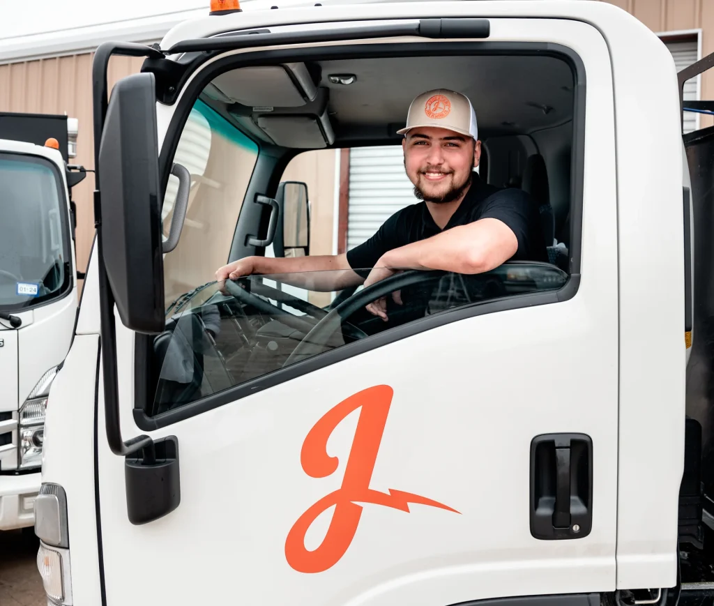 Happy driver in a cap smiles from a white truck featuring an orange J-bolt logo.