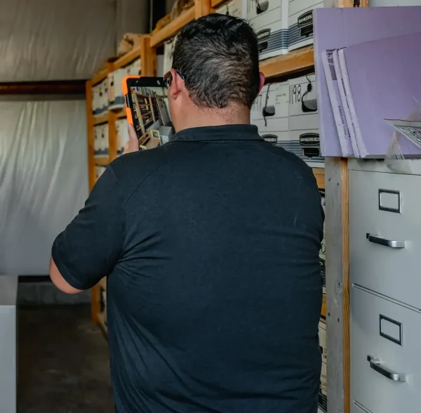 Man photographing archival boxes and animal skulls with a tablet in a storage area. TITLE: