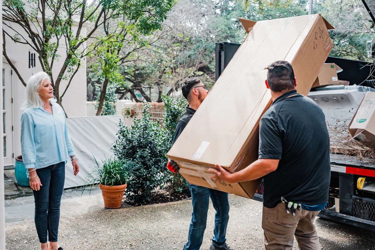 Two movers carry a large cardboard box while a woman watches in a sunny outdoor setting. TITLE