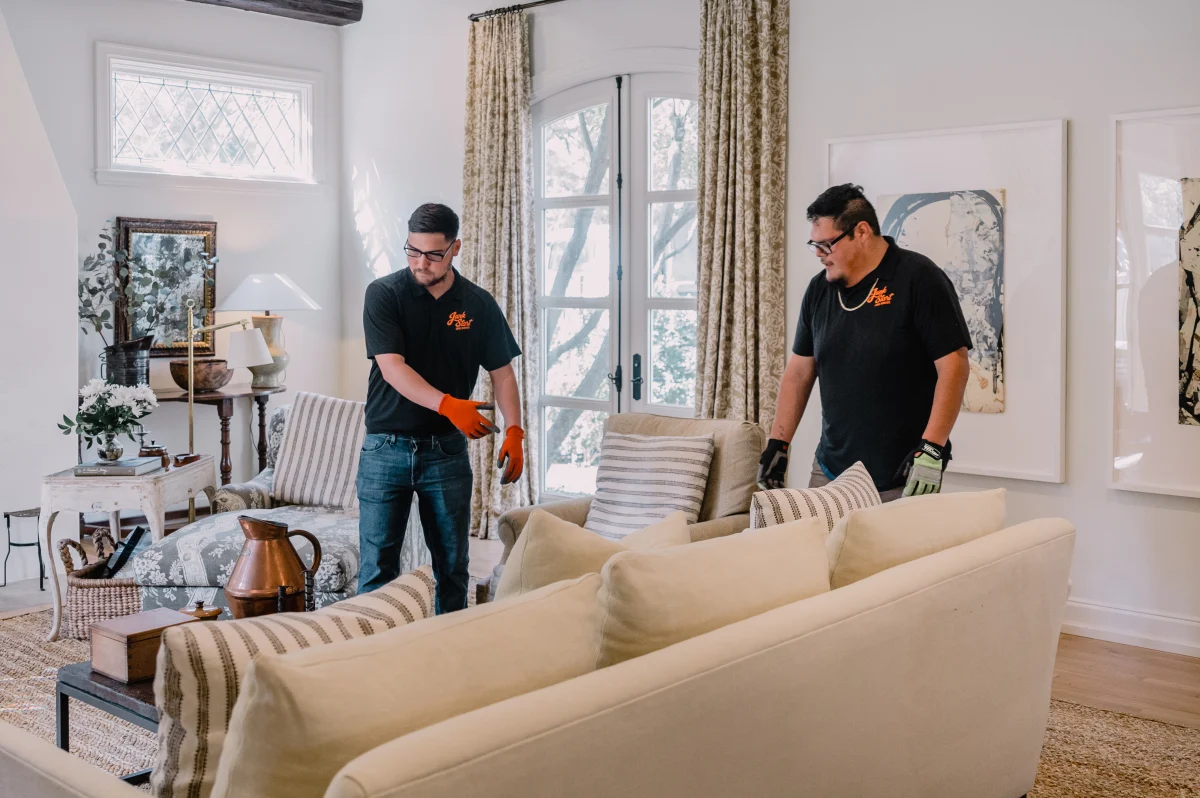 Two men in Junk Start shirts and gloves carefully move a light-colored sofa in a