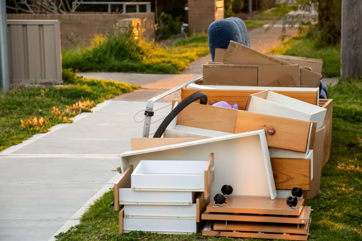 Disassembled furniture, cardboard boxes, and a bicycle tire are piled on a sunny lawn next to