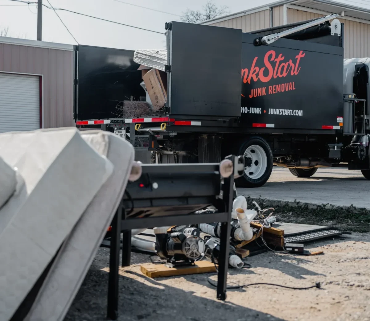 A black JunkStart junk removal truck is loaded with debris as mattresses and items sit nearby